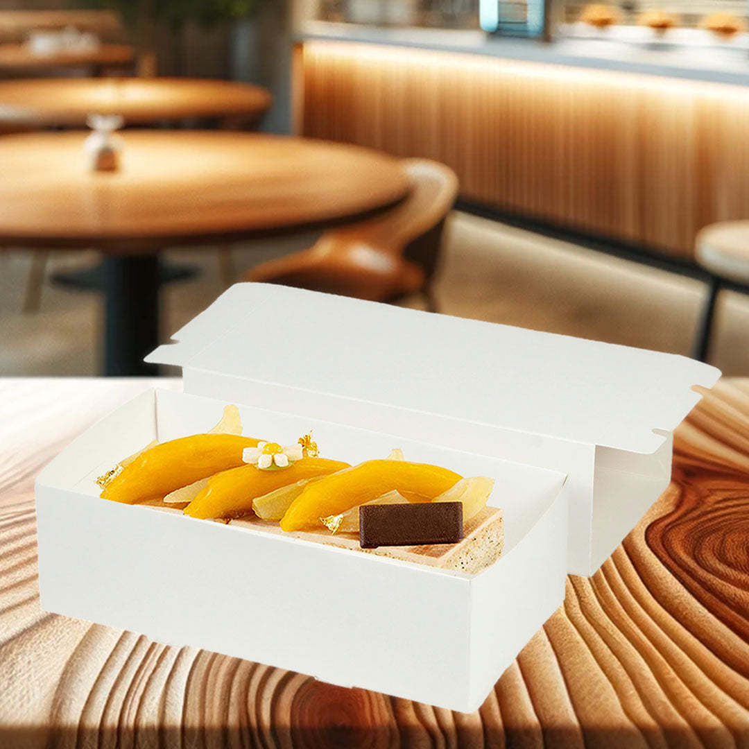 La imagen muestra una caja de cartón blanca abierta, dentro de la cual se exhibe un elegante postre, biodegradable, ideal para empacar postres, de fondo una pastelería elegante