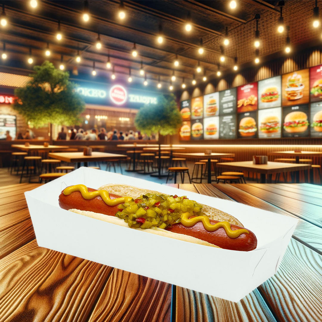 Charola de cartón blanca biodegradable diseñada para servir hot dogs, mostrando un hot dog dentro. El fondo muestra un restaurante de comida rápida