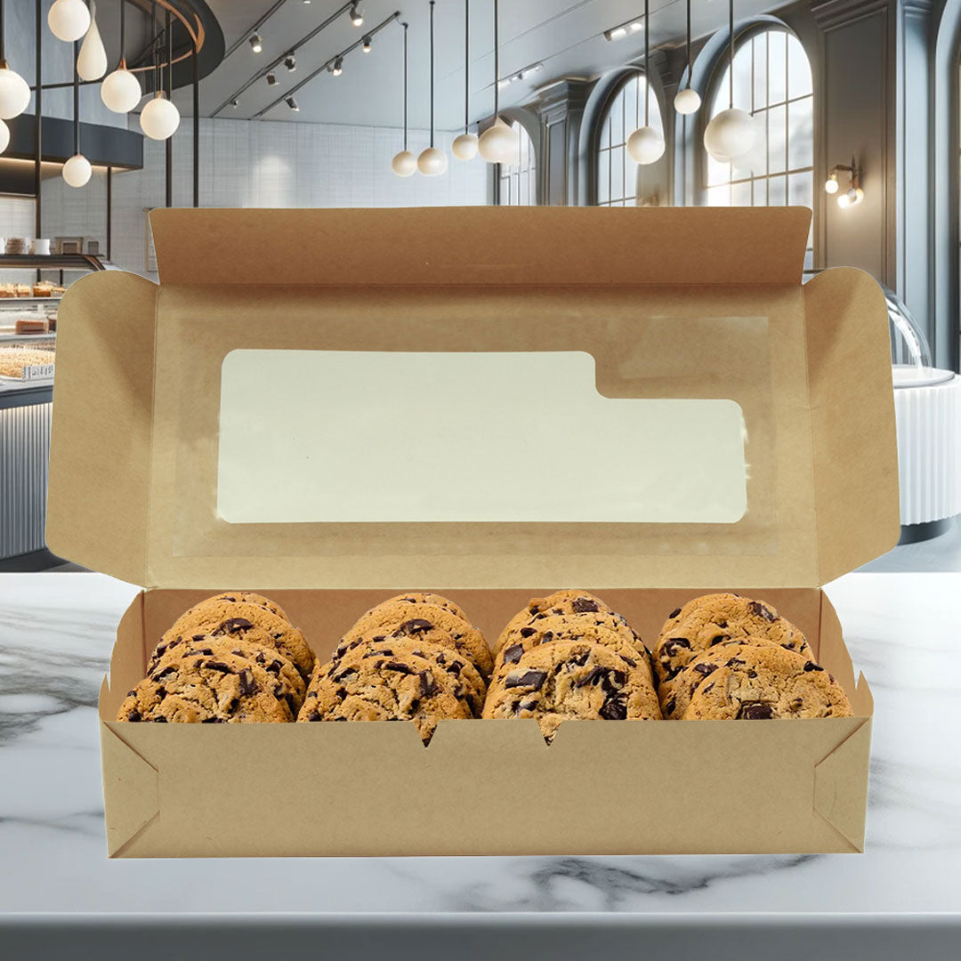 La imagen muestra una caja rectangular de cartón kraft biodegradable, abierta. En el interior de la caja hay seis galletas. La tapa tiene una ventana transparente de plástico, La caja está ubicada en una pastelería. El diseño de la caja es resistente